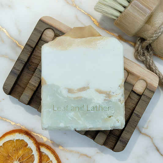 Bar of soap with 'Leaf and Lather' branding on a wooden soap dish with dried oranges and a brush.