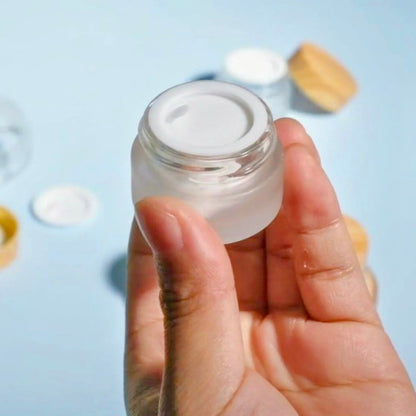 Hand holding a small glass jar with a white lid against a light blue background
