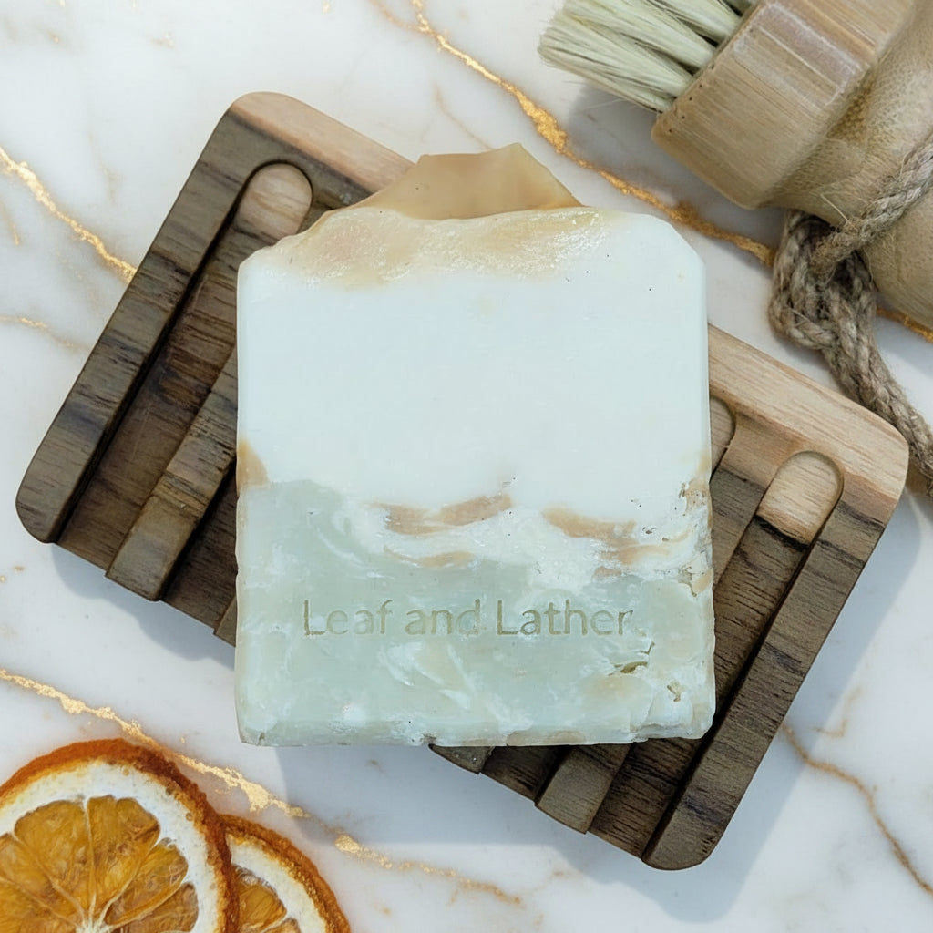 Bar of soap with 'Leaf and Lather' branding on a wooden soap dish with dried oranges and a brush.