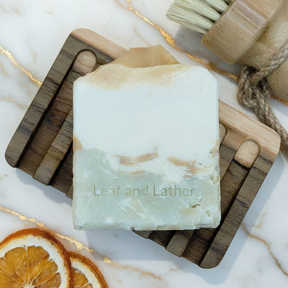 Bar of soap with 'Leaf and Lather' branding on a wooden soap dish with dried oranges and a brush.