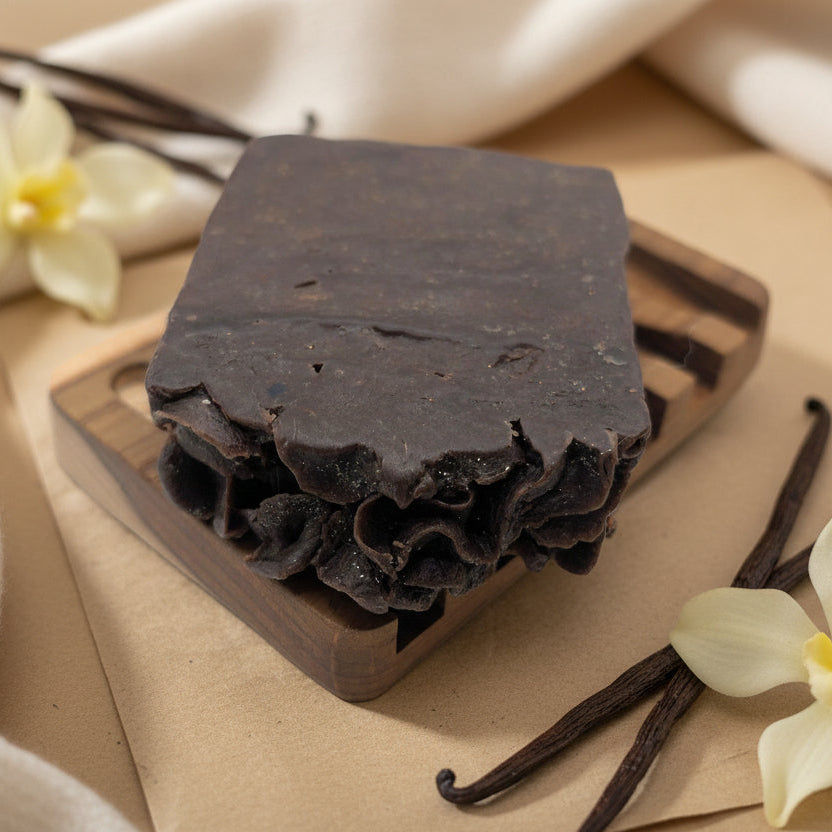 Dark soap bar on a wooden board with vanilla beans and flowers on a beige background