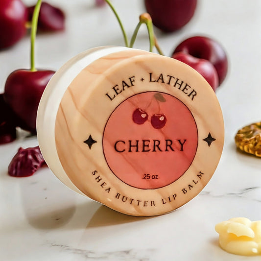 Jar of Leaf + Lather Cherry Shea Butter Lip Balm on a marble surface with cherries in the background.