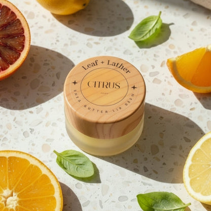 Jar of 'Leaf + Lather' Citrus lip balm surrounded by citrus fruits on a textured surface